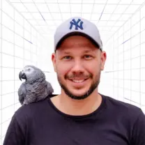 Man wearing a gray NY cap smiles with an African grey parrot perched on his shoulder in a white grid backdrop.